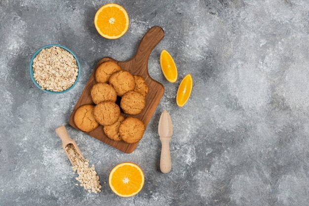 Groothoek foto van zelfgemaakte koekjes op een houten bord en havermout met sinaasappelen over grijs oppervlak.