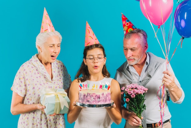 Gratis foto grootouders die verjaardagsgiften houden dichtbij meisje met cake blazende kaarsen op blauwe achtergrond