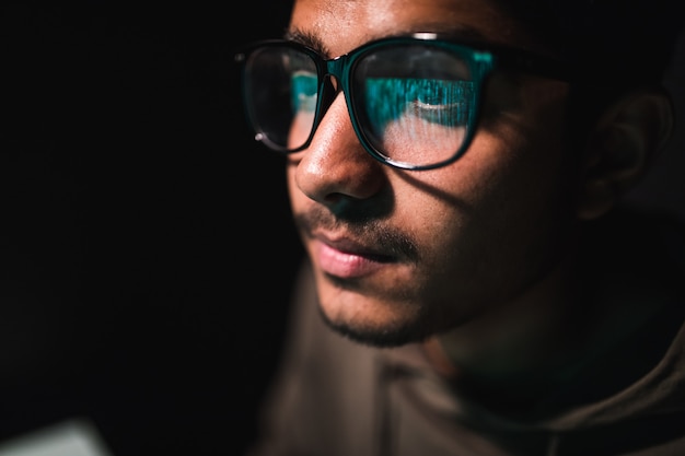 Hacker in bril en een kap werkt op een computer in het donker, een weerspiegeling in een bril
