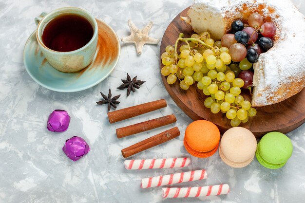 Half-bovenaanzicht suikerpoeder cake met verse druiven macarons kaneel en thee op witte ondergrond fruit cake koekje zoete suiker bakken