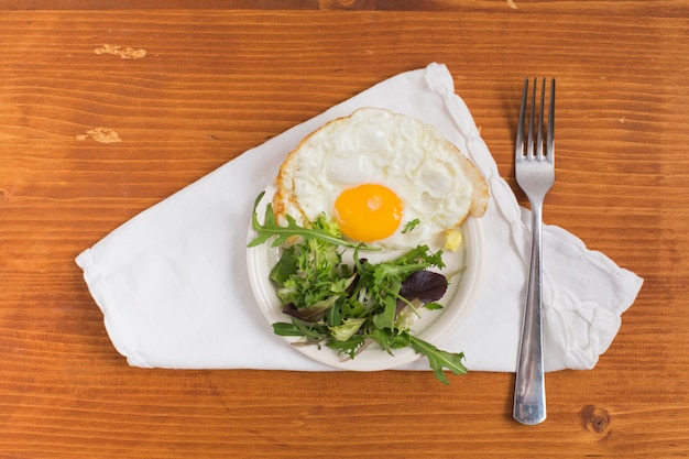 Half gefrituurd ei met salade op plaat en vork over het witte servet tegen houten geweven achtergrond