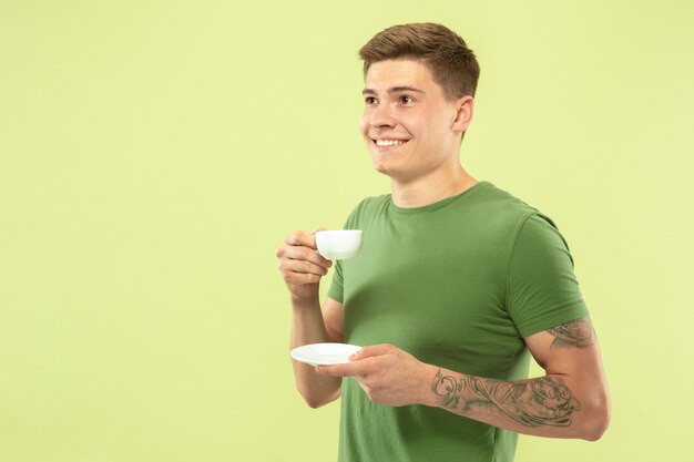 Half-lengteportret van de Kaukasische jonge man op groene studioachtergrond. Mooi mannelijk model in overhemd. Concept van menselijke emoties, gezichtsuitdrukking, verkoop, advertentie. Genieten van koffie of thee drinken.