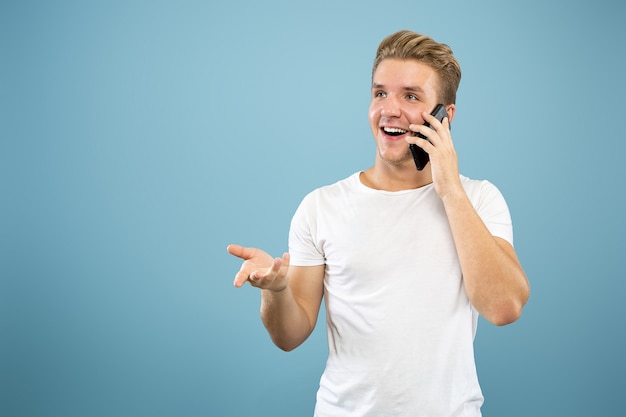 Half-length portret van een blanke jonge man op blauwe studio achtergrond. Mooi mannelijk model in overhemd. Concept van menselijke emoties, gezichtsuitdrukking, verkoop, advertentie. Praten aan de telefoon, ziet er blij uit.