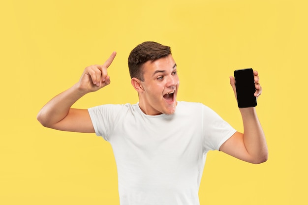 Half-length portret van een blanke jonge man op gele studio achtergrond. Mooi mannelijk model in overhemd. Concept van menselijke emoties, gezichtsuitdrukking, verkoop, advertentie. Toont het scherm van de telefoon en glimlacht.
