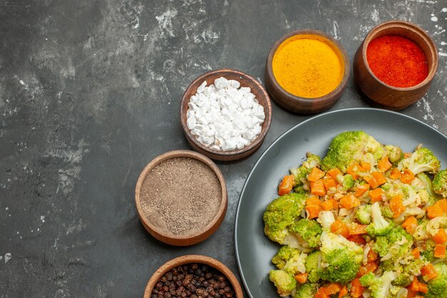 Half schot van gezonde maaltijd met brocoli en wortelen op een zwarte plaat en kruiden op grijze tafel