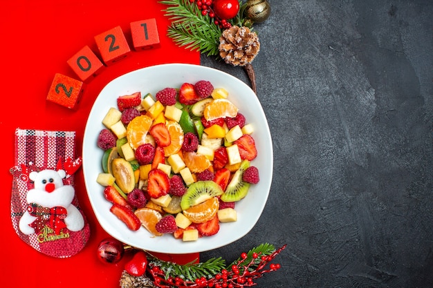 Half schot van verzameling van vers fruit op diner plaat decoratie accessoires fir takken en cijfers op een rood servet aan de rechterkant op donkere achtergrond