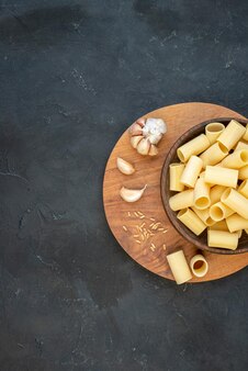 Half shot rauwe pasta's in een bruine kom knoflookrijst op ronde houten plank aan de linkerkant op zwarte achtergrond