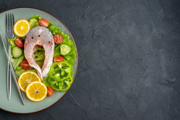 Half shot rauwe vis en verse groenten, schijfjes citroen en bestek op een grijze plaat aan de linkerkant op een zwarte ondergrond met vrije ruimte