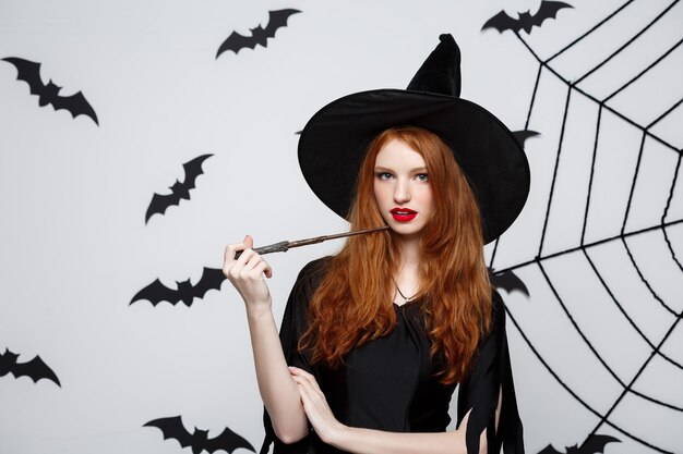 Halloween-concept - mooie heks die met magische stok op grijze muur speelt.
