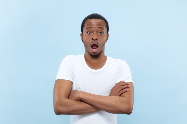 Halve lengte close-up portret van jonge Afro-Amerikaanse man in wit overhemd op blauwe achtergrond. Menselijke emoties, gezichtsuitdrukking, advertentie, verkoop, concept. Staande handen gekruist, geschokt en verbaasd.