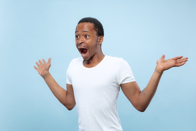 Halve lengte close-up portret van jonge Afro-Amerikaanse man in wit overhemd op blauwe achtergrond. Menselijke emoties, gezichtsuitdrukking, advertentie, verkoopconcept. Uitnodigend, ziet er geschokt en verbaasd uit. Copyspace.