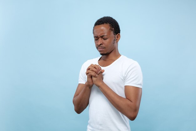 Halve lengte close-up portret van jonge Afro-Amerikaanse man in wit overhemd op blauwe achtergrond. Menselijke emoties, gezichtsuitdrukking, advertentieconcept. Bidden met gesloten ogen, ziet er hoopvol uit.