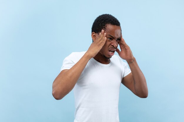 Halve lengte close-up portret van jonge Afro-Amerikaanse man in wit overhemd op blauwe achtergrond. Menselijke emoties, gezichtsuitdrukking, advertentieconcept. Lijdend aan hoofdpijn, zware gedachten, mentale problemen.