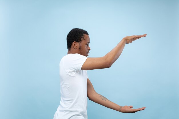 Halve lengte close-up portret van jonge Afro-Amerikaanse man in wit overhemd op blauwe muur. Menselijke emoties, gezichtsuitdrukking, advertentieconcept. Houd een lege balk vast, copyspace voor uw tekst.