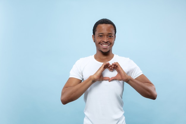 Halve lengte close-up portret van jonge Afro-Amerikaanse man in wit overhemd op blauwe muur. Menselijke emoties, gezichtsuitdrukking, advertentieconcept. Met het teken van een hart door zijn handen, glimlachend.