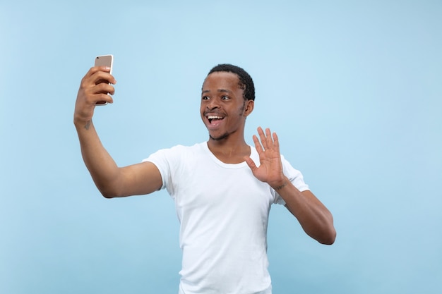 Halve lengte close-up portret van jonge Afro-Amerikaanse man in wit overhemd op blauwe ruimte. Menselijke emoties, gezichtsuitdrukking, advertentie, concept