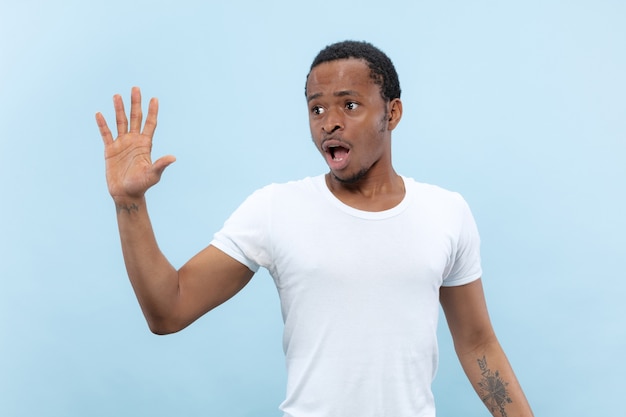 Halve lengte close-up portret van jonge Afro-Amerikaanse man in wit overhemd op blauwe ruimte. Menselijke emoties, gezichtsuitdrukking, advertentie, verkoopconcept