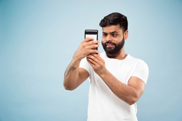 Halve lengte close-up portret van jonge hindoo man in wit overhemd op blauwe achtergrond. Menselijke emoties, gezichtsuitdrukking, advertentieconcept. Negatieve ruimte. Selfie of videoblog, vlog, chating maken.