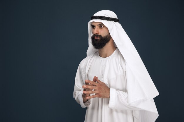 Halve lengte portret van Arabische Saoedische zakenman op donkerblauwe studioachtergrond. Jong mannelijk model staat en ziet er attent uit. Concept van zaken, financiën, gezichtsuitdrukking, menselijke emoties.