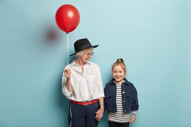 Halve lengte shot van aanhankelijke oma feliciteert klein kind met de eerste dag op school, houdt rode ballon met blije uitdrukkingen. Vrolijke oma, kleindochter komt in een goed humeur terug van circusvoorstelling