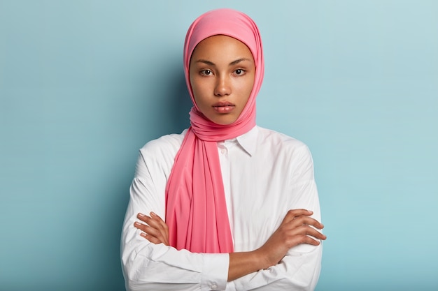 Halve lengte shot van ernstige zelfverzekerde vrouw draagt traditionele roze hijab, houdt de armen gekruist, gekleed in een wit overhemd, staat tegen de blauwe muur, luistert met belangstelling naar de gesprekspartner. Religie