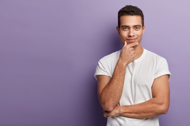 Halve lengte shot van knappe Europese man houdt kin, kijkt met tevreden zelfverzekerde uitdrukking direct, draagt wit t-shirt, poseert over paarse muur, vrije ruimte opzij