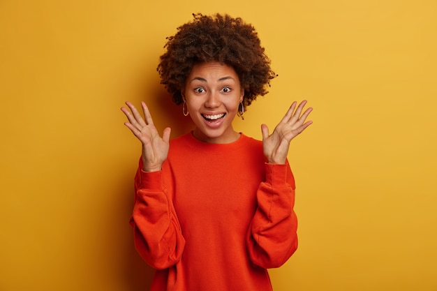 Halve lengte shot van optimistische gekrulde vrouw houdt handpalmen omhoog, gekleed in een rode trui, voelt zich opgewonden door uitstekend nieuws te horen, glimlacht breed, staat tegen een gele achtergrond.