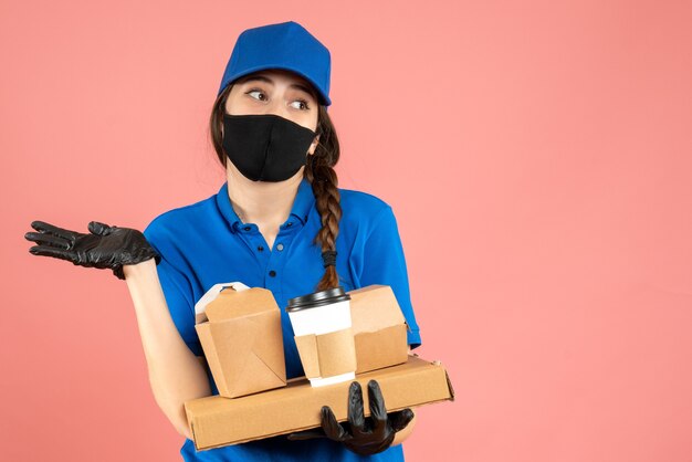 Halve lichaamsopname van een denkend koeriersmeisje met een medisch masker en handschoenen met bestellingen op een pastelkleurige perzikachtergrond