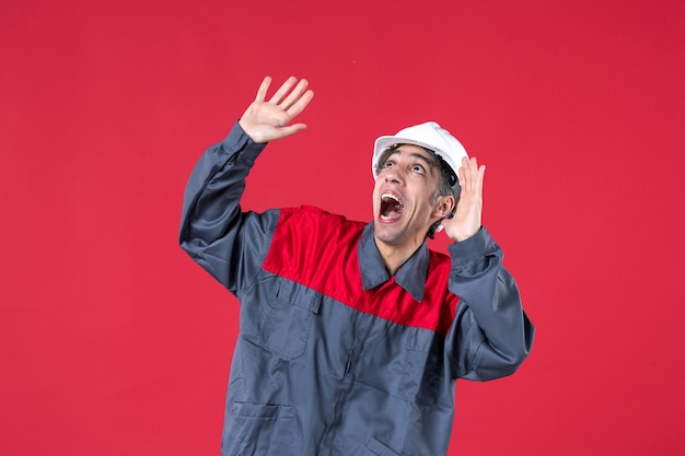 Halve lichaamsopname van een nerveuze jonge bouwer in uniform met een helm die omhoog kijkt op een geïsoleerde rode muur