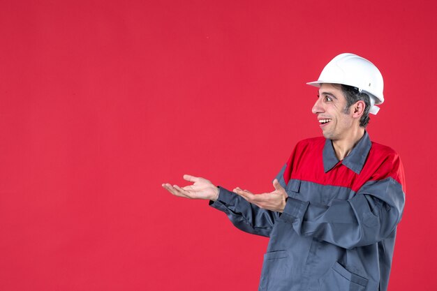 Halve lichaamsopname van lachende nieuwsgierige jonge architect in uniform met helm op geïsoleerde rode muur