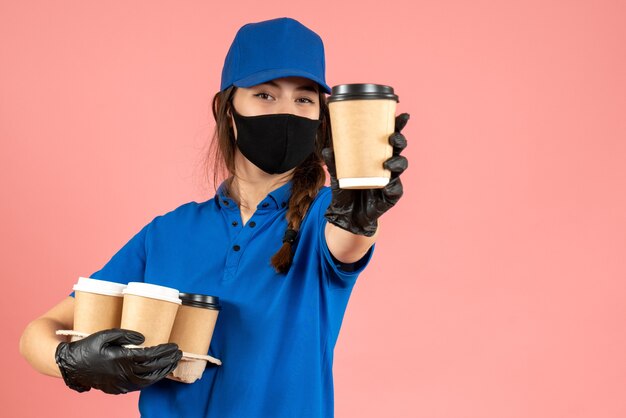 Halve lichaamsopname van zelfverzekerd koeriersmeisje met zwarte medische maskerhandschoenen met koffie op pastelkleurige perzikachtergrond