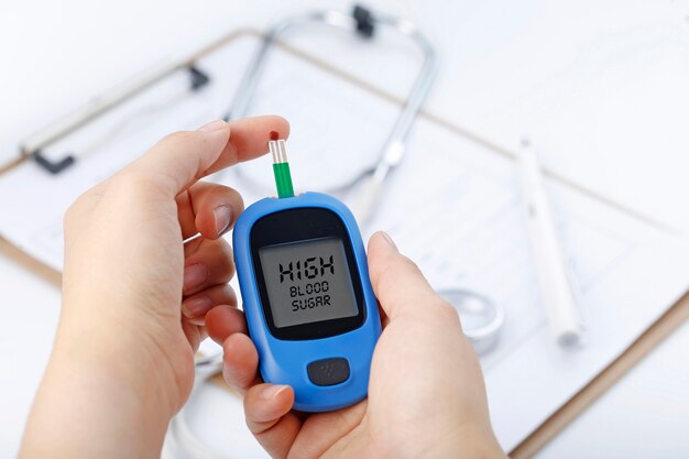 Hand met een bloedglucosemeter met bloedglucose, de achtergrond is een stethoscoop en een grafisch bestand