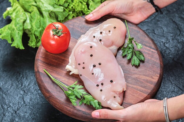 Hand met houten plaat van rauwe kipfilet met Groenen op donkere tafel.