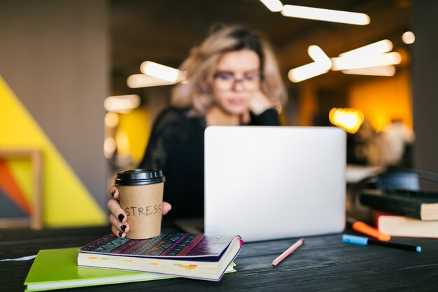 Handen van jonge mooie vrouwenzitting bij lijst in zwart overhemd die aan laptop in mede-werkend bureau, bezige student freelancer werken, die koffie drinken