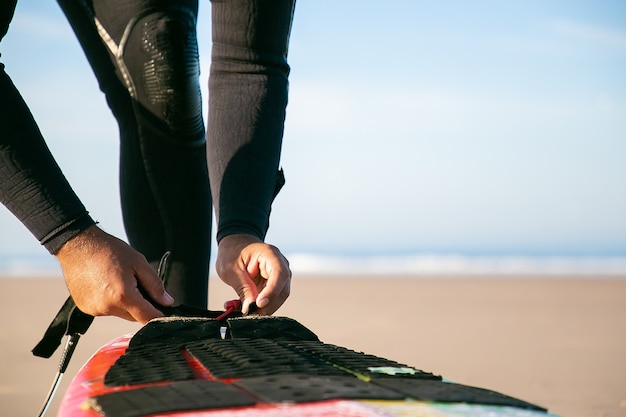 Handen van surfer in wetsuit die surfplank aan zijn enkel op oceaanstrand binden