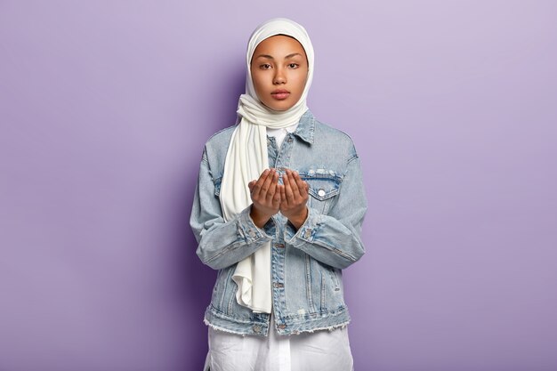 Handgebaar en biddende concept. Ernstige vrouw met donkere huid steekt handen in gebed, pleit voor iets, draagt sjaal en spijkerjasje, geïsoleerd over paarse muur. Moslim religie concept