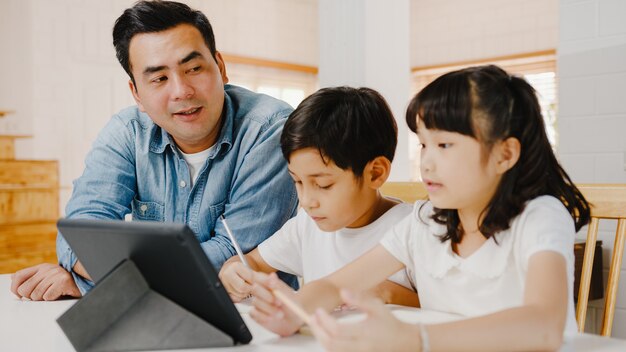 Happy Asia family homeschooling, vader leert kinderen thuis met behulp van digitale tablet in de woonkamer.