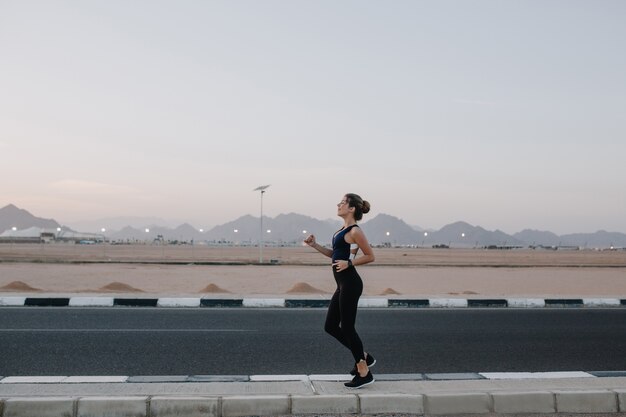 Hardlopen, trainen op de weg in de vroege ochtend van vrolijke mooie vrouw. Opleiding van een sterke sportvrouw, energie, motivatie, gezonde levensstijl, opgewekte stemming.