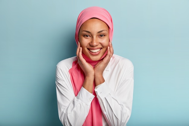 Headshot van aangenaam uitziende moslimvrouw raakt wangen met beide handen aan, toont witte tanden, draagt wit overhemd en roze sluier, geïsoleerd tegen blauwe muur, drukt vreugde, geluk, verrukking uit
