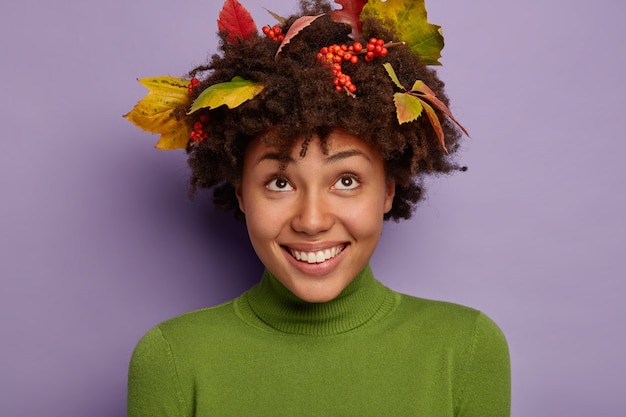 Headshot van aangenaam uitziende vrolijke vrouw heeft brede glimlach, kijkt boven, verheugt zich met mooi kapsel versierd met herfstbladeren