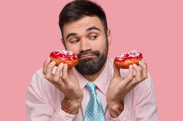 Headshot van aantrekkelijke jonge man heeft gezichtsuitdrukking verbaasd, houdt smakelijke donuts vast, ziet er verwarrend uit, heeft verleiding