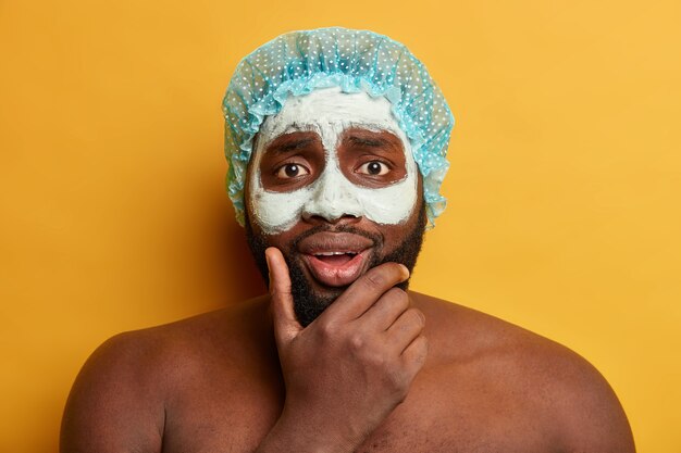 Headshot van Afro man houdt kin vast, past klei-moddermasker toe voor een gezonde huid, kijkt met bezorgde uitdrukking, draagt een douchemuts