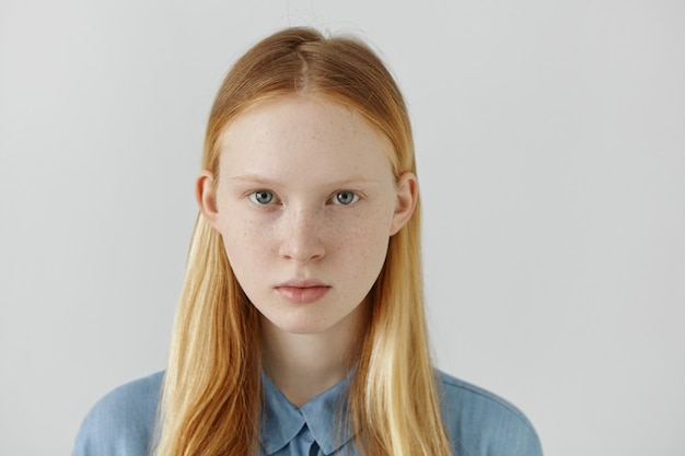 Headshot van blanke meisje met sproeten, blond haar en lichte ogen gekleed in blauwe school shirt staande tegen de witte muur