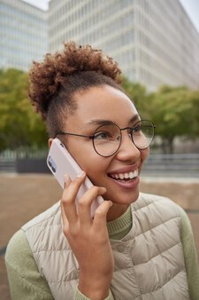 Headshot van een gelukkige vrouw met krullend haar geniet van een telefoongesprek glimlacht positief geconcentreerd in de verte draagt een ronde bril gekleed in vrijetijdskleding