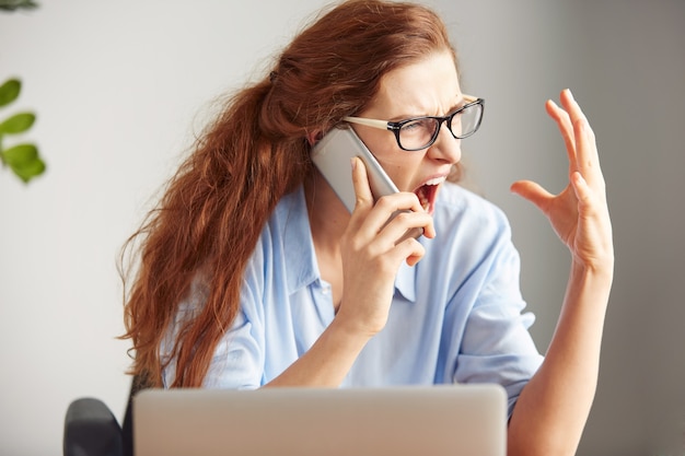 Headshot van een jonge vrouwelijke baas die van woede schreeuwt op de mobiele telefoon terwijl hij aan het bureau zit