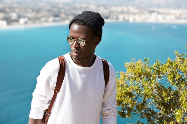 Headshot van ernstige afrikaanse man tegen het schilderachtige uitzicht op de europese zeehaven stad. reiziger in stijlvolle kleding en zonnebril die peinzend en verward op zoek is naar een overnachting