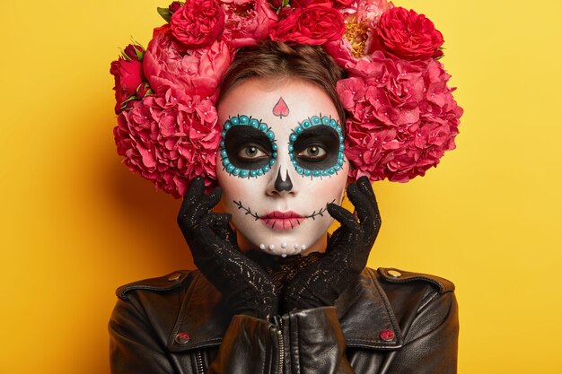 Headshot van ernstige mooie vrouw met schedelmake-up, gezicht geschilderd door kunstenaar, draagt zwarte kleding, wil er spookachtig uitzien, poseert tegen gele achtergrond. Traditionele Mexicaanse feestdag