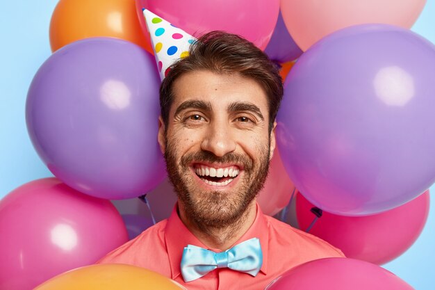 Headshot van gelukkig Europese man draagt papieren kegel hoed, roze shirt en vlinderdas, ziet er positief uit