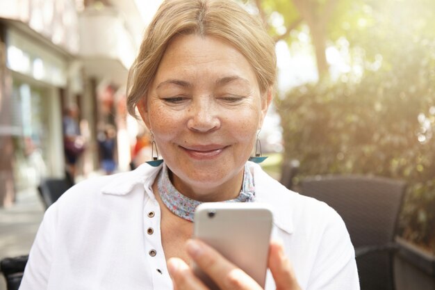 Headshot van gelukkige oude blonde vrouw met blond haar en mooie glimlach kijken naar het scherm van haar elektronische gadget, communiceren met haar kinderen online met behulp van slimme telefoon, zittend op café buitenshuis