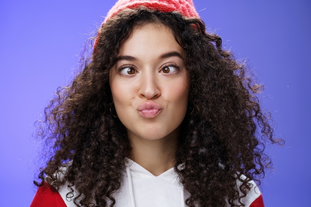 Headshot van grappig en speels meisje dat voor de gek houdt en gezichten maakt die niet bang zijn om grappige lippen te vouwen zoals eend en loensend ijs, kinderachtig en hilarisch te apen over blauwe muur in wintermuts.